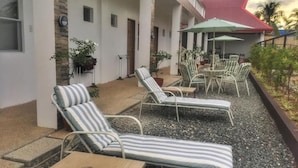 Un espace extérieur aménagé, abrité et doté de chaises longues et de tables, entouré d’un bâtiment blanc avec un toit rouge.