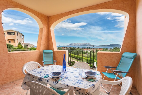 Un balcon avec une table dressée pour deux personnes, des chaises et une vue sur une montagne et la mer, qui se dressent à l’horizon.