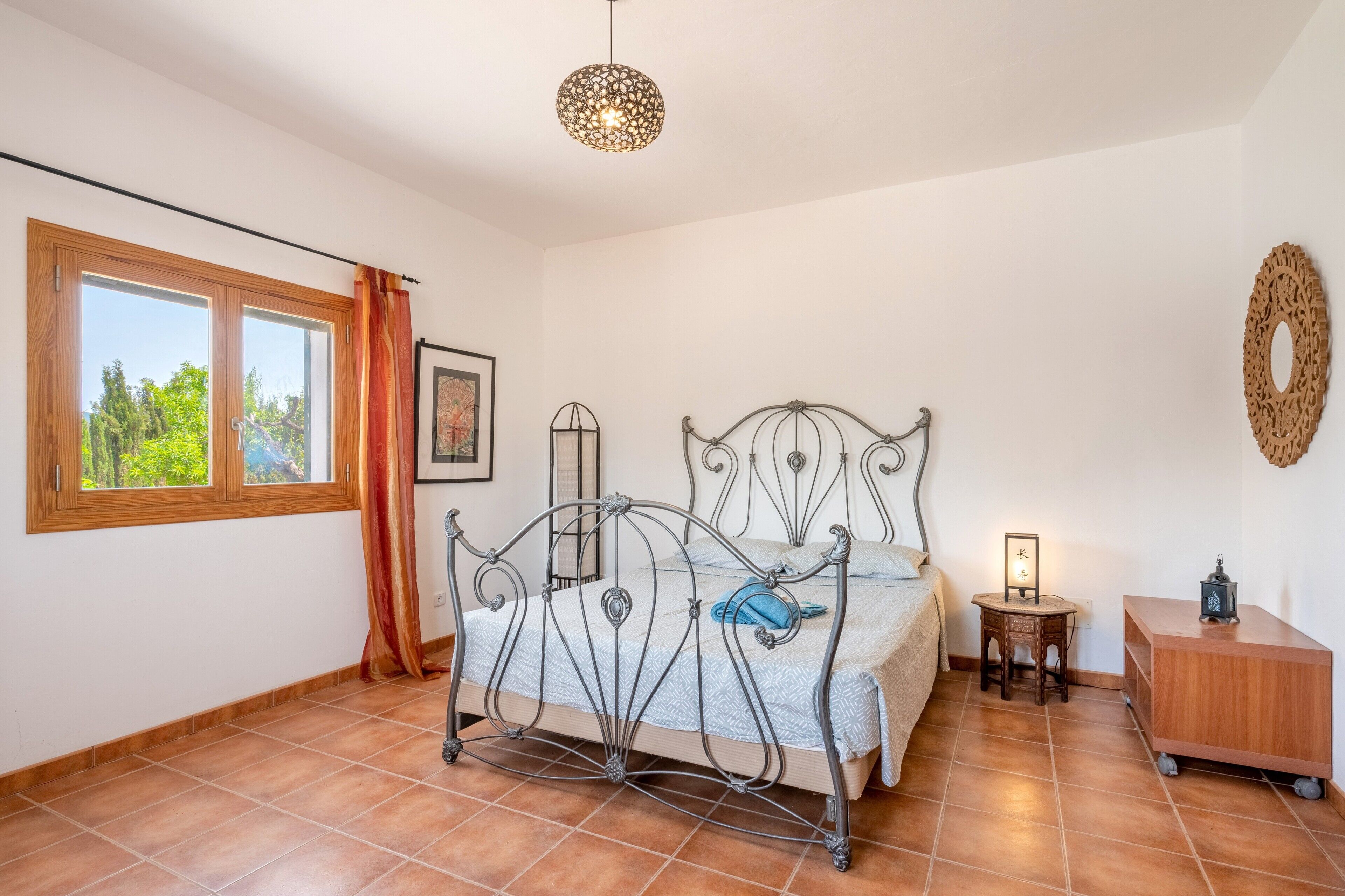 A bedroom with a metal bed frame, a bedside table with a lamp, a small wooden cabinet, and a window with curtains.