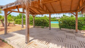 Eine Hollaube mit einem Reetdach, umgeben von einer Holzterrasse, Palmen und einem Pool im Hintergrund.