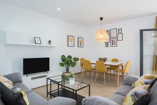 A modern living room with a flat-screen TV, sofa, coffee table, dining area with yellow chairs, and a potted plant. - Beautiful Villa Sandra (just families) with Pool, Air Conditioning & Terraces (Cádiz)