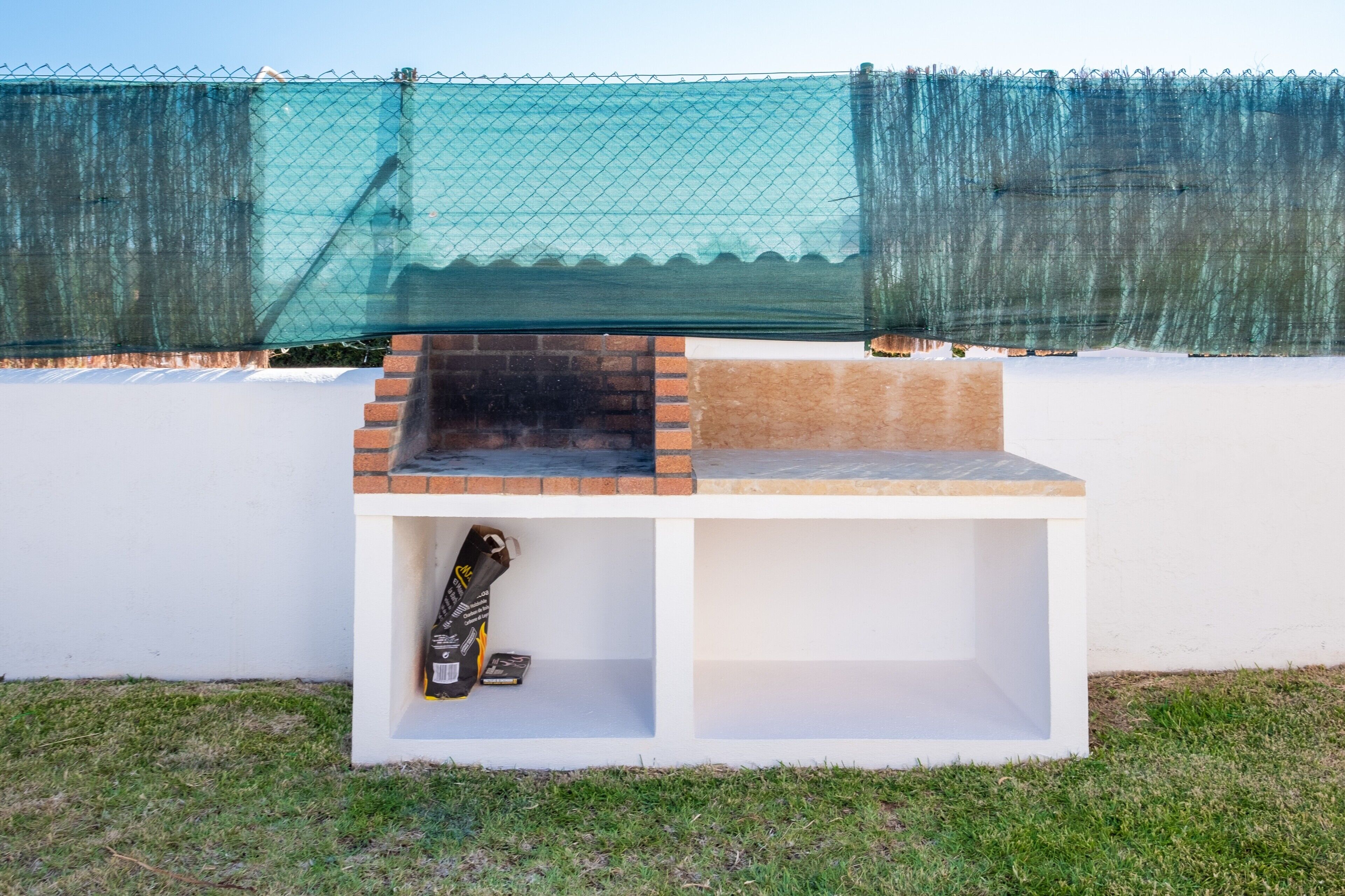 Una pequeña zona de parrilla al aire libre con base de ladrillo y repisa blanca, ubicada frente a una pared blanca con una cerca de malla verde al fondo.