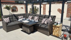 Une terrasse extérieure avec un canapé d’angle, une table dressée pour les repas et une pile de bois de chauffage.