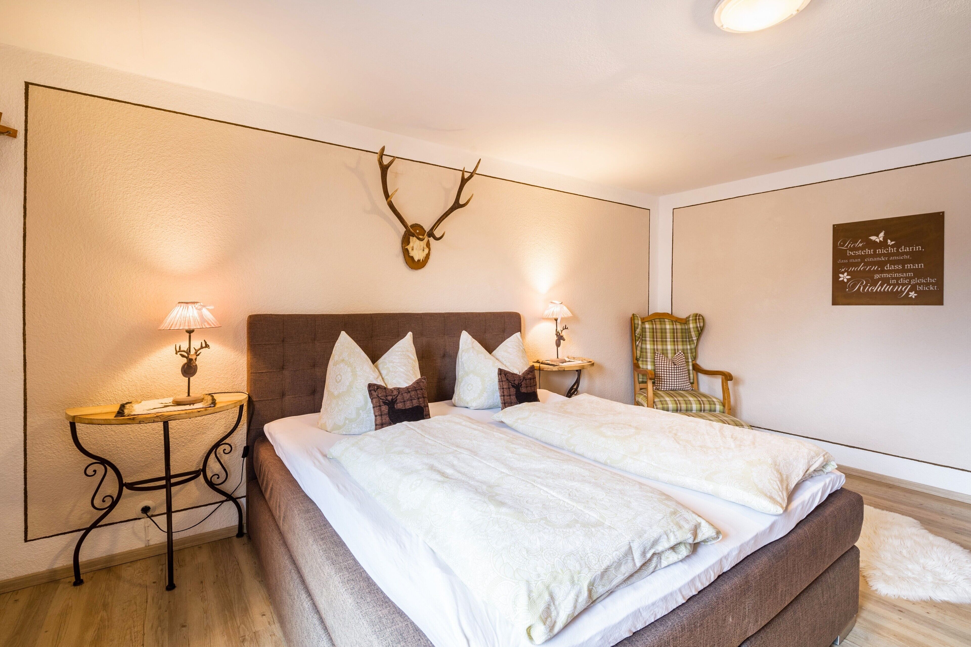 Une chambre à coucher avec un grand lit, deux tables de chevet avec des lampes, une chaise et une décoration murale représentant un crâne de cerf.