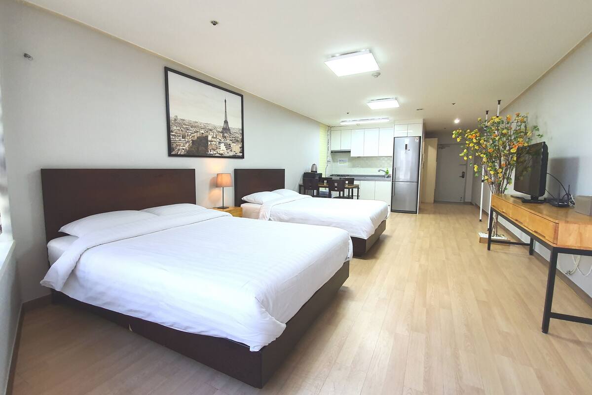 Una habitación de hotel con dos camas, una kitchenette pequeña, un comedor y un televisor sobre una mesa de madera.
