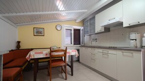 A compact kitchen with white cabinets, a dining table with chairs, a clock, and a window with blinds. - Apartment Slunjski for 4 people (Njivice)