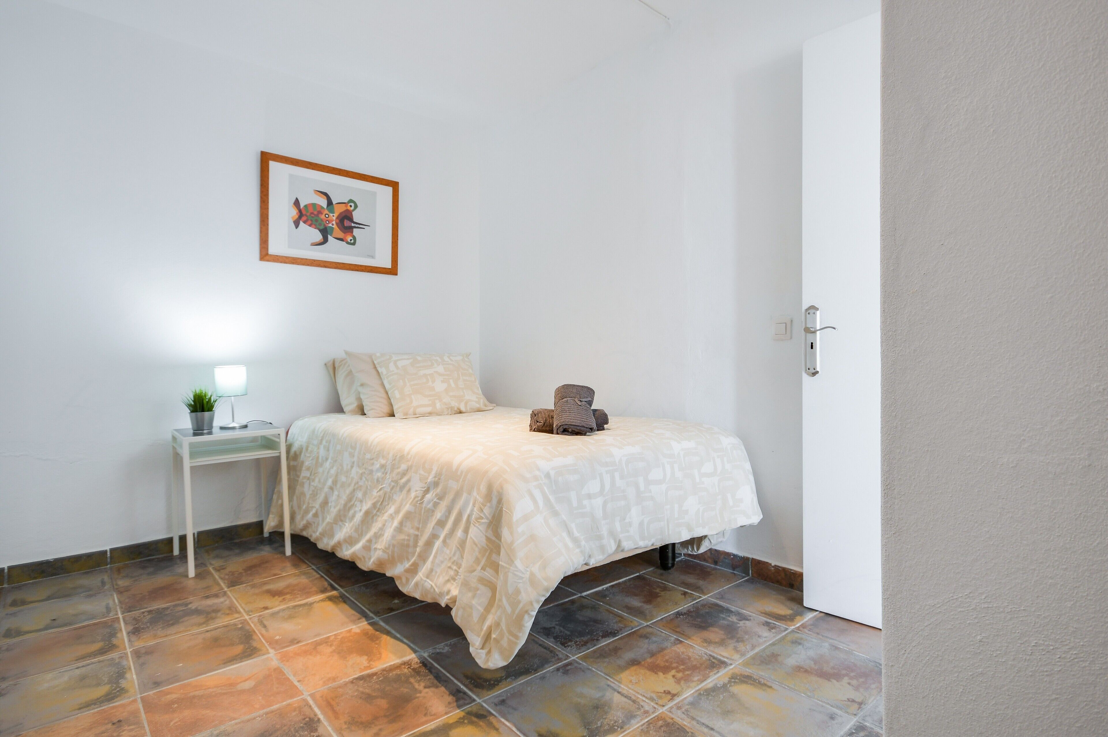 A bedroom with a bed, a bedside table with a lamp, a framed picture on the wall, and a door.