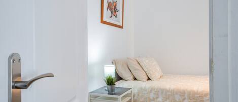 Un dormitori amb un llit, una tauleta de nit amb una làmpada, un quadre al mur i una porta amb maneta.