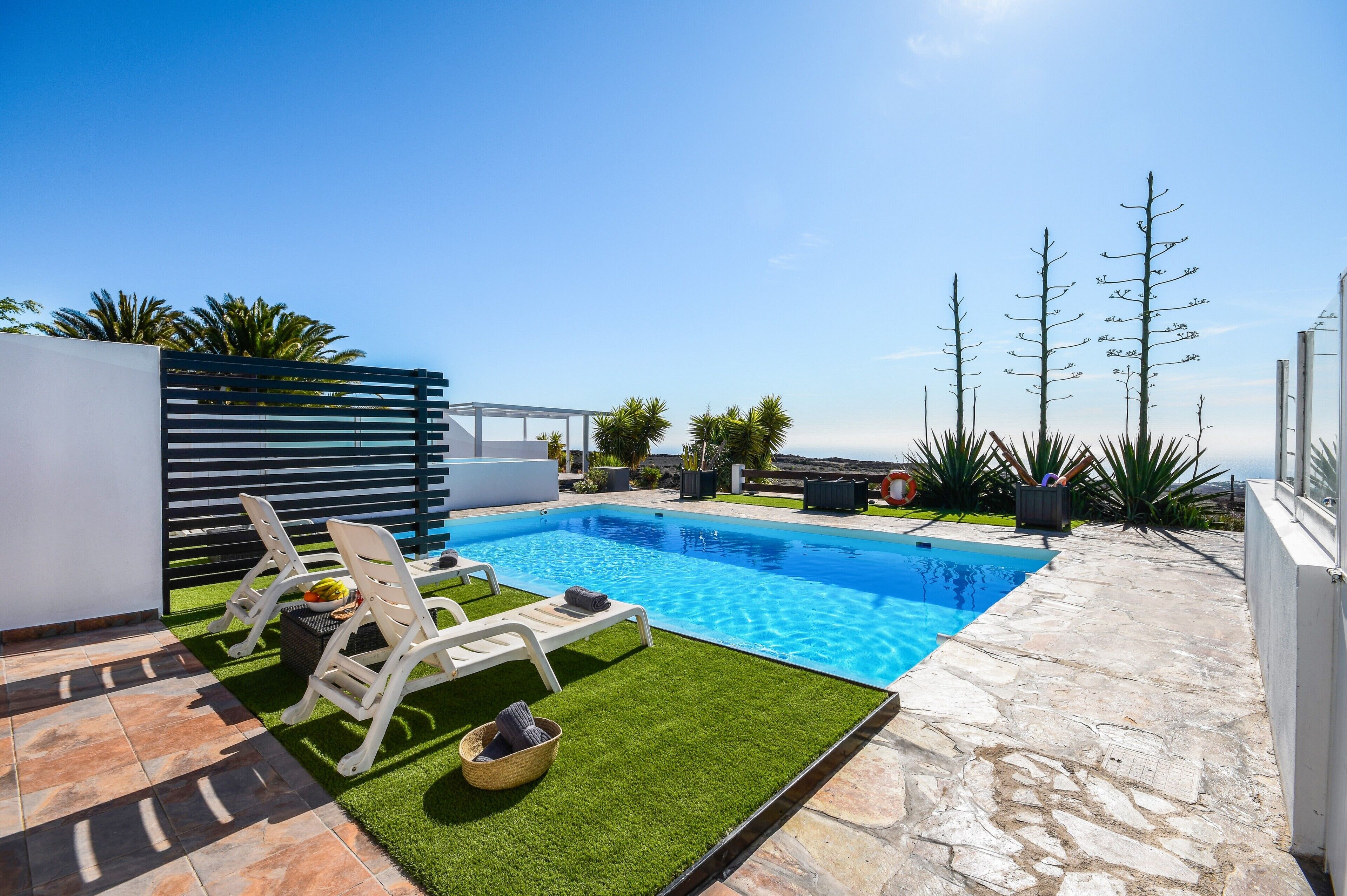 Uma piscina com deck de pedra, espreguiçadeiras brancas e vista para o mar.