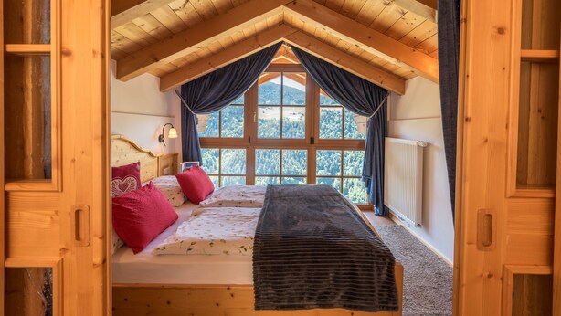 Een slaapkamer met een houten plafond, een bed met rode kussens, een raam met gordijnen en een radiator.