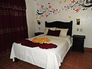 A neatly made bed with a white and maroon bedspread, a yellow towel, and a red throw. There are two bedside tables with lamps, a decorative wall with floral patterns, and a red curtain.