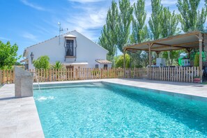 Una piscina con solárium de madera, una zona de estar cubierta y un edificio blanco con tejado de azulejos.