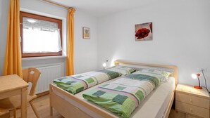 Un dormitorio con cama, escritorio, silla, mesita de noche con lámpara, ventana con cortinas y un cuadro en la pared.