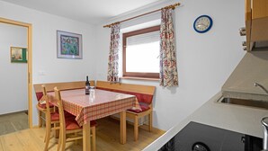Una zona de comedor con una mesa, sillas, un banco, una ventana con cortinas, un reloj de pared y un lavamanos de cocina.