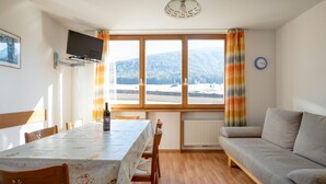 Una habitación con una mesa de comedor, sillas, un sofá, un televisor montado en la pared, una ventana con cortinas y un reloj.