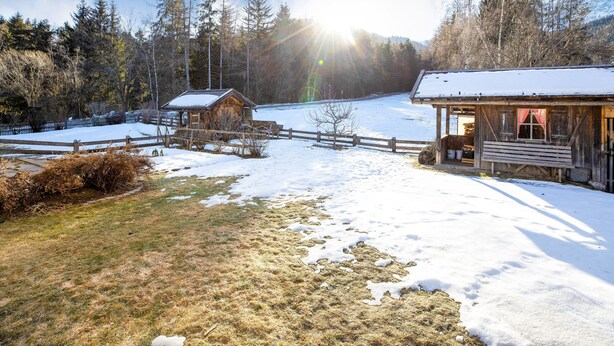 A wooden cabin with a snow-covered roof, a wooden fence, and a clear view of a forested mountain range in the background.