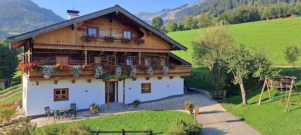 A wooden chalet with a balcony, a patio with outdoor seating, and a view of a green valley.