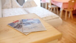 A neatly arranged hotel room with a bed, a wooden table with magazines, a dining table with chairs, and a window with curtains. - Holiday Apartment “Kruma - Gsail 2” with Mountain View, Wi-Fi & Balcony (Olang)