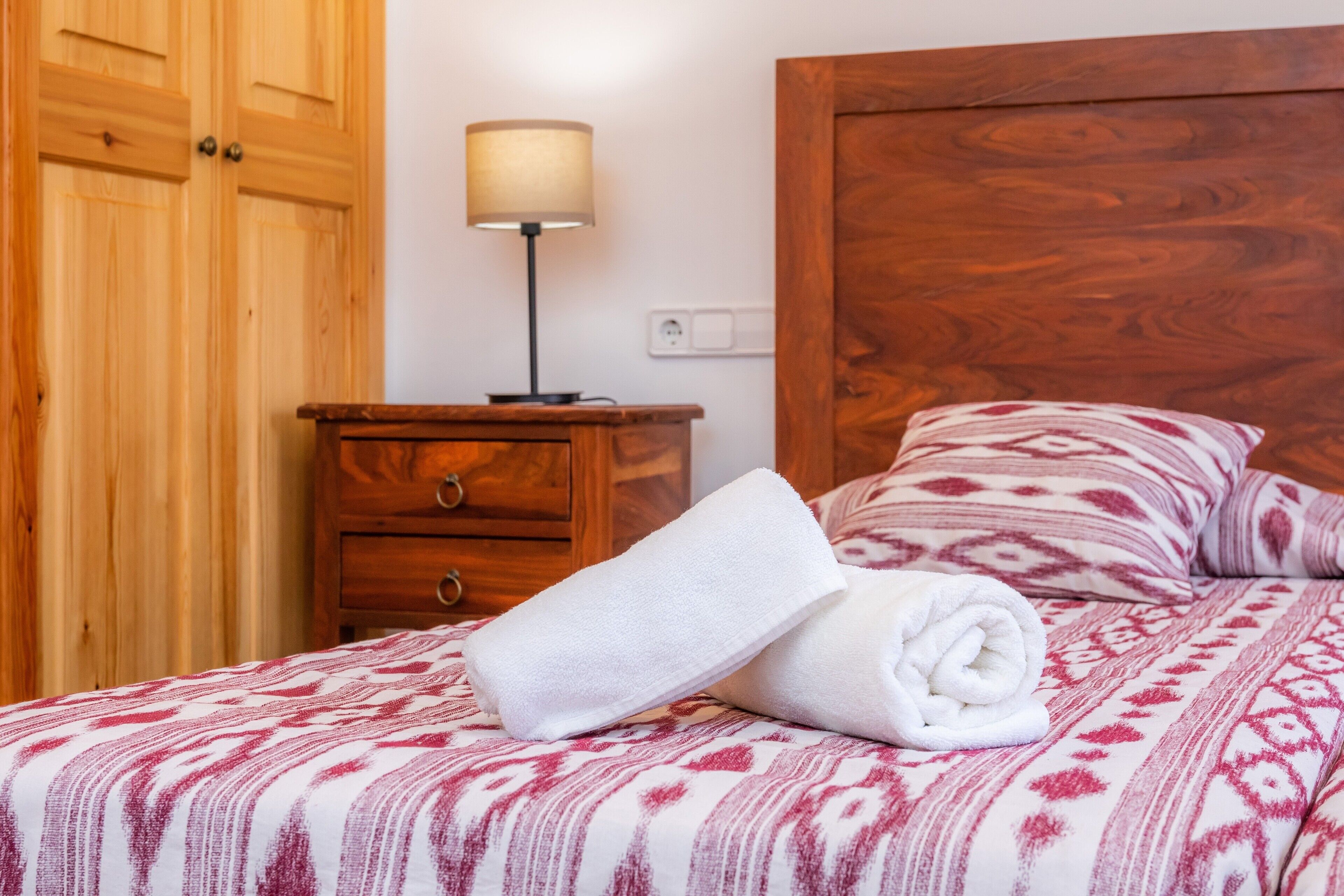 Una cama bien hecha con una colcha estampada, una mesita de noche de madera con una lámpara y un armario de madera.