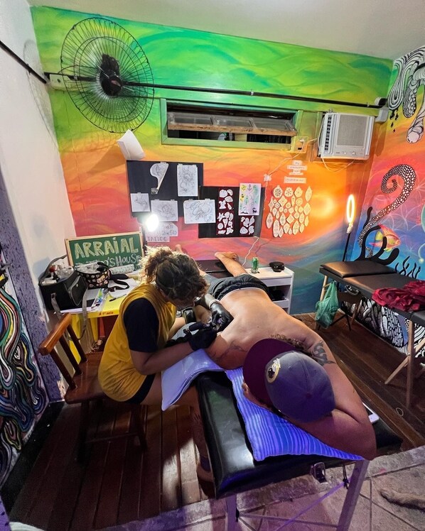 A tattoo parlor with a colorful mural, a fan, and various tattoo designs on the wall. - Chamos Hostel Cultural (Arraial do Cabo)