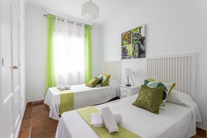 A hotel room with two beds, a window with green curtains, a radiator, and a wall-mounted artwork.