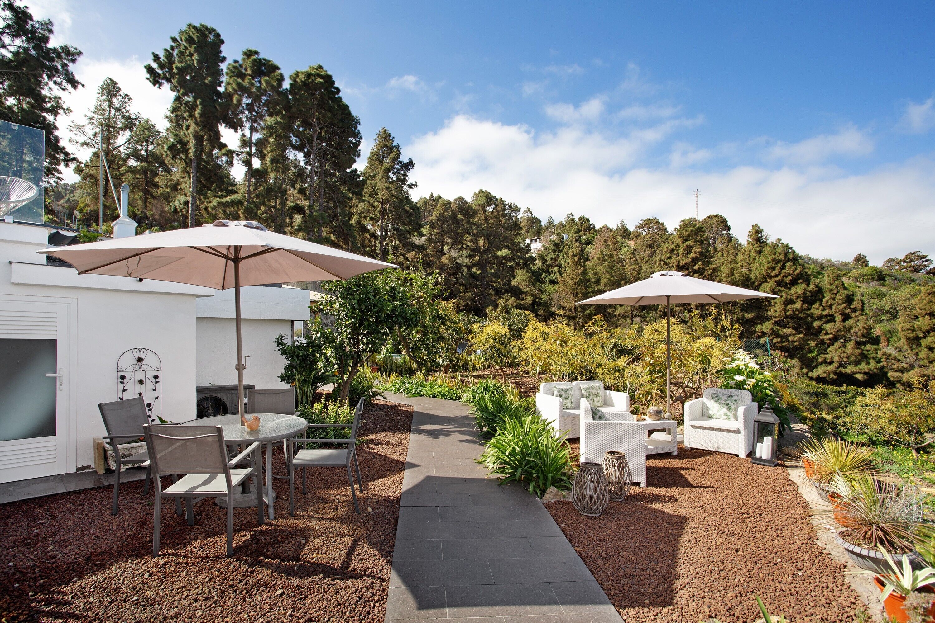 A patio area with outdoor furniture, two umbrellas, and a pathway leading to a white building.