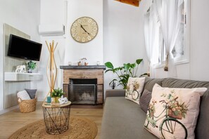 Un salon avec une cheminée, une horloge, un téléviseur et un canapé agrémenté de coussins décoratifs.