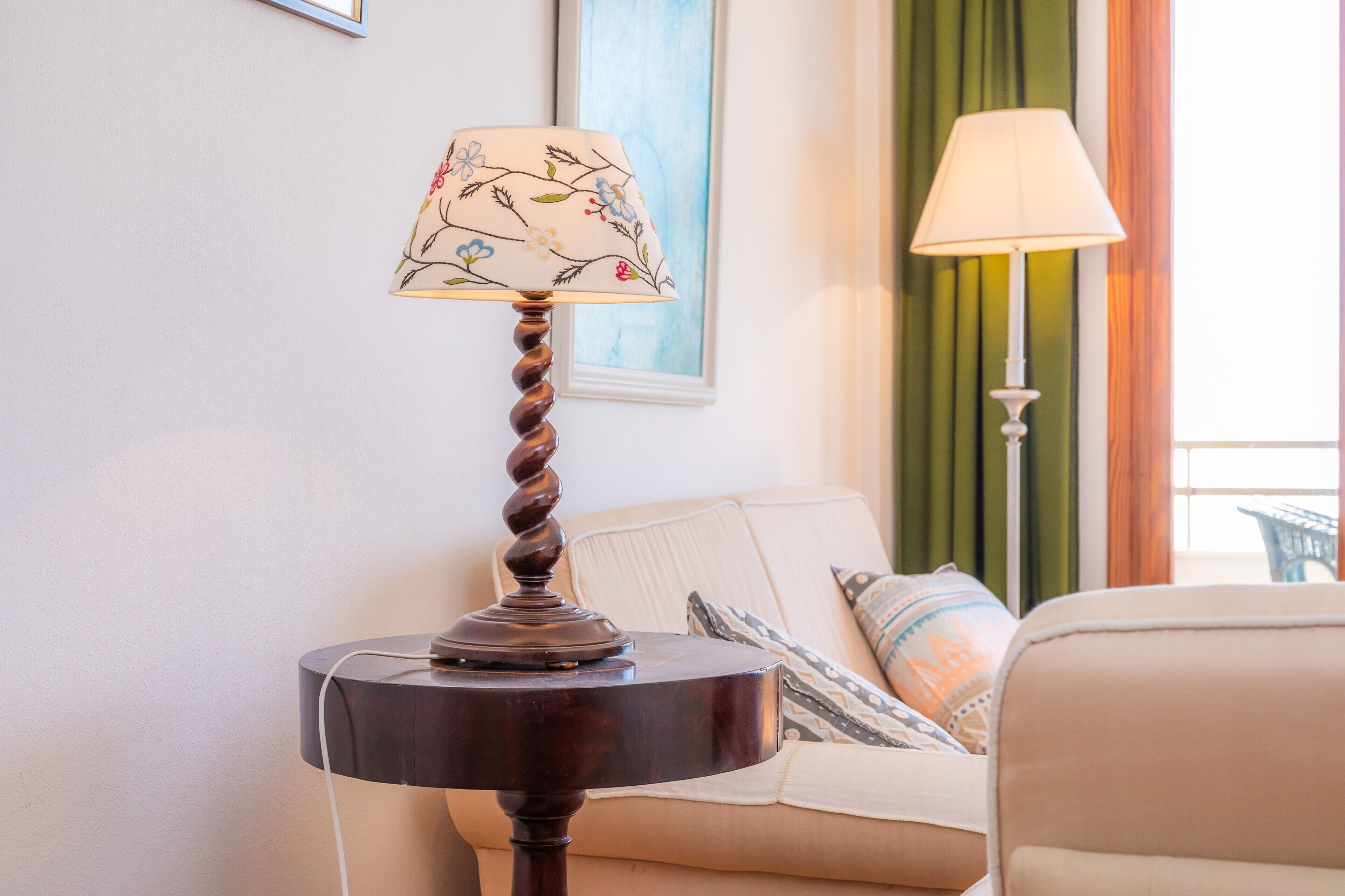 A round table with a lamp, a sofa with patterned cushions, and a painting on the wall.