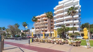 A multi-story building with balconies, outdoor seating with yellow umbrellas, and palm trees. -  Central Apartment at the Beach Promenade with Wi-Fi and Sea View (Cala Millor)
