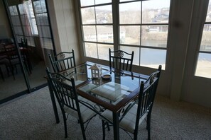 A dining area with a glass-top table surrounded by four chairs, a small decorative item on the table, and a view of a body of water and buildings through large windows.