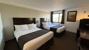 A hotel room with two beds, a desk, a lamp, a window with curtains, and a painting on the wall.