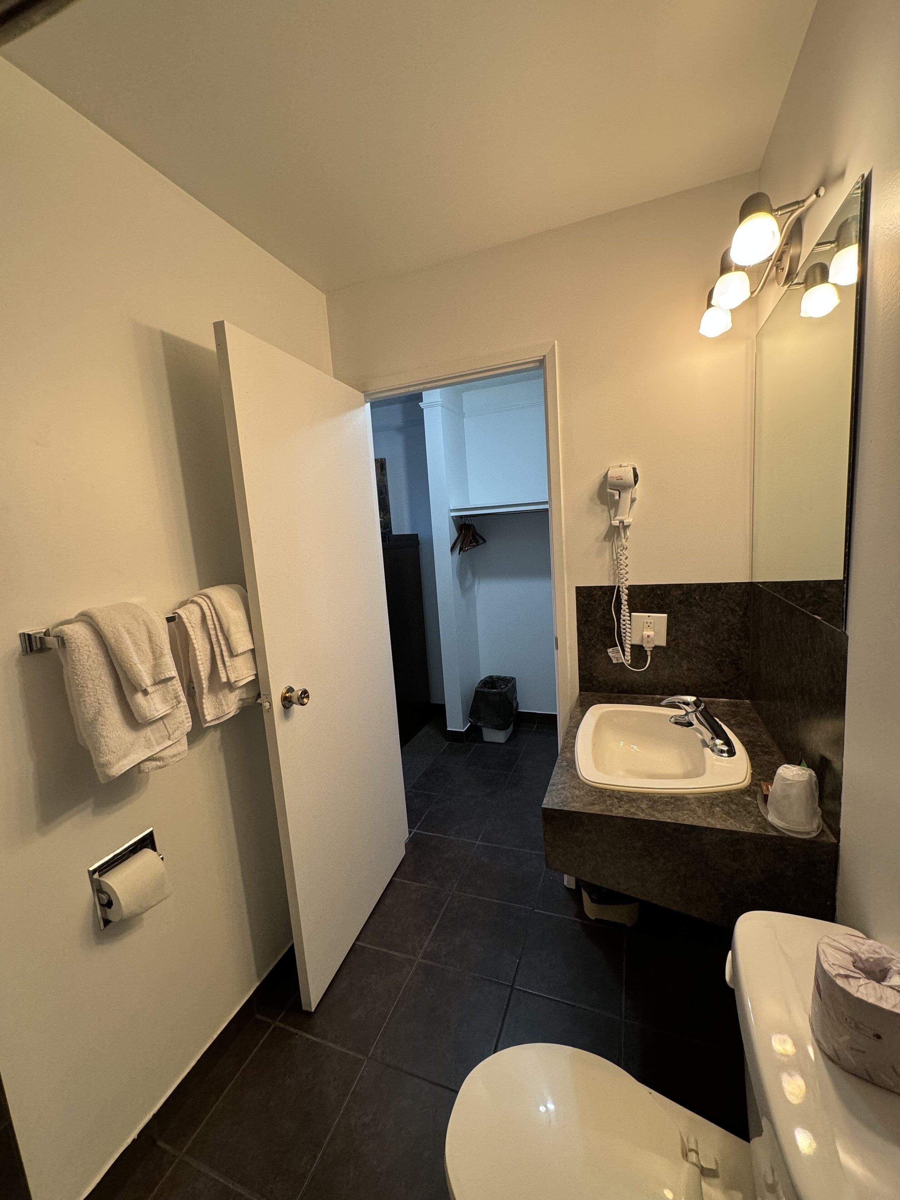 A bathroom with a toilet, sink, and mirror. There is a towel rack with towels, a trash can, and a door leading to another room.