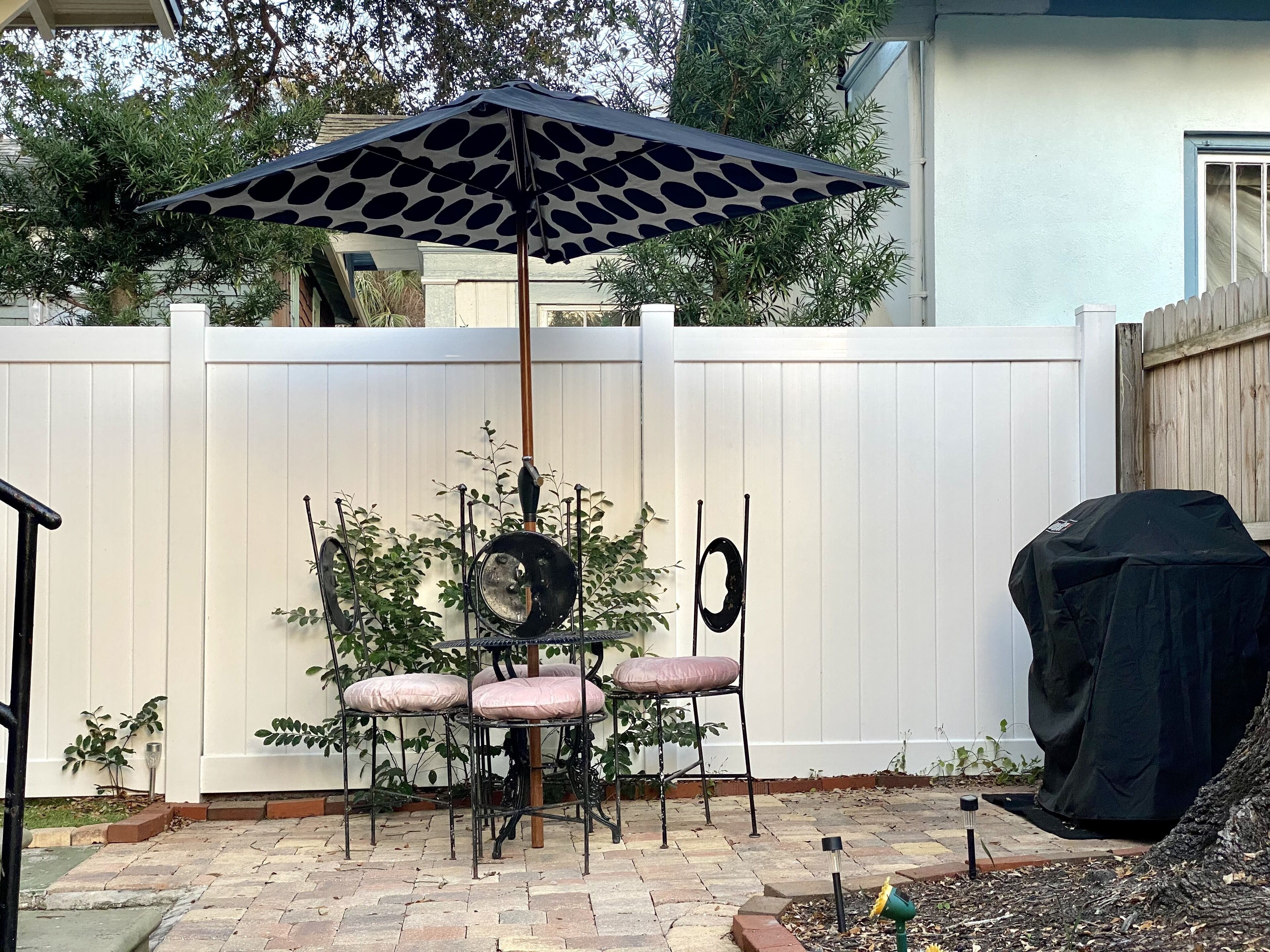 A patio with a table and chairs under a patterned umbrella, a barbecue grill, and a white fence.