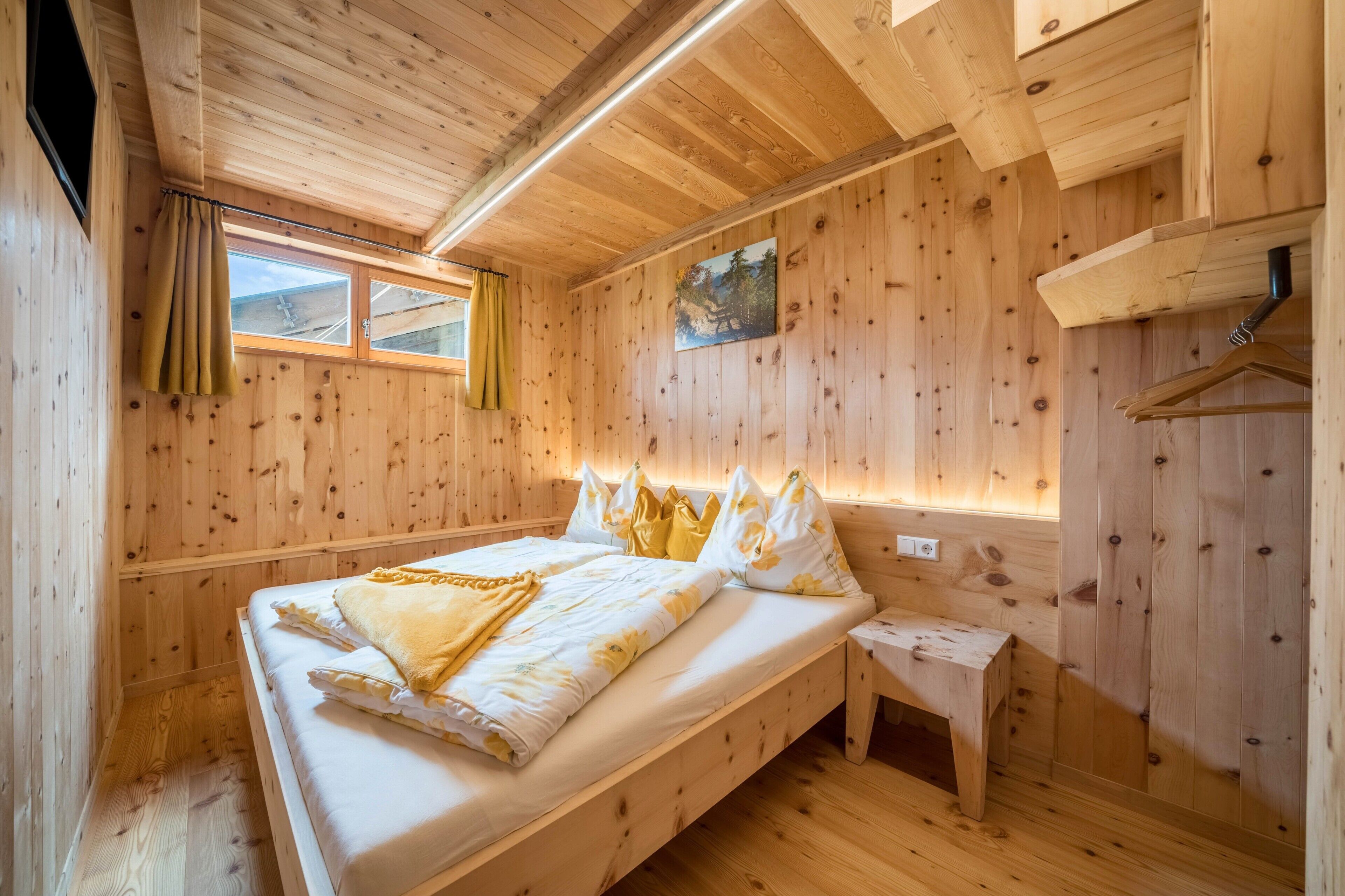 A wooden cabin room with a bed, a bench, a window, and a painting on the wall.