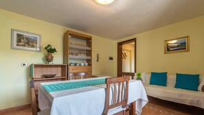 A dining area with a table set for two, a wooden chair, a sofa with blue cushions, and a shelf with glassware. - Vacation Home “Chia B” Close to Beach with Sea View & Garden (Chia)