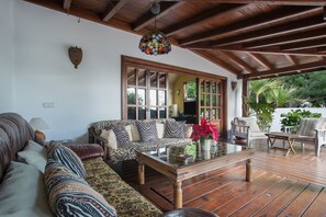 A room with a wooden ceiling, a sofa with patterned cushions, a glass coffee table, and a view of greenery outside.