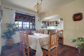 A dining area with a table set for four, wooden chairs, a chandelier, a potted plant, and a view into the kitchen.