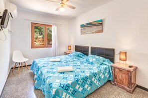 Une chambre à coucher comprenant un lit, une chaise, une table de chevet, un ventilateur de plafond, une fenêtre et un tableau accroché au mur.