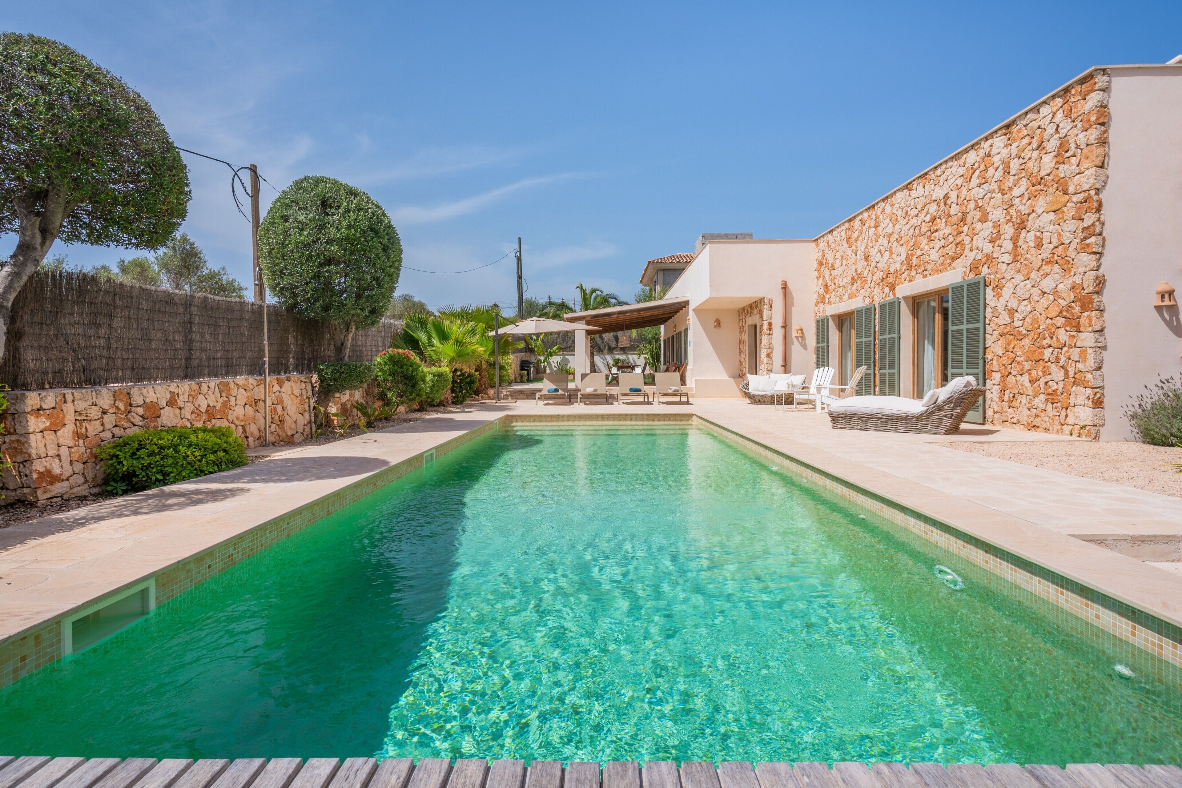 Une piscine avec un mur en pierre d’un côté et un patio couvert équipé de chaises longues.
