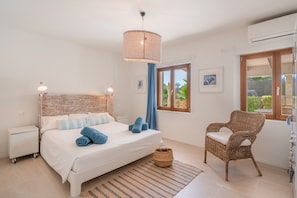Un dormitorio con cama, una silla de mimbre, una mesita de noche, una ventana con cortinas y un aire acondicionado instalado en la pared.