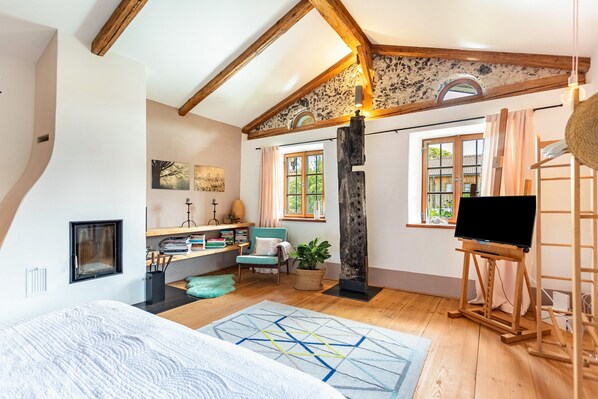 A bedroom with a sloped ceiling, wooden beams, a bed, a desk, a chair, a potted plant, a painting, a window, and a television.