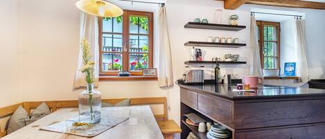 A kitchen with wooden beams, a dining table, a hanging light fixture, and a window with a view of flowers.