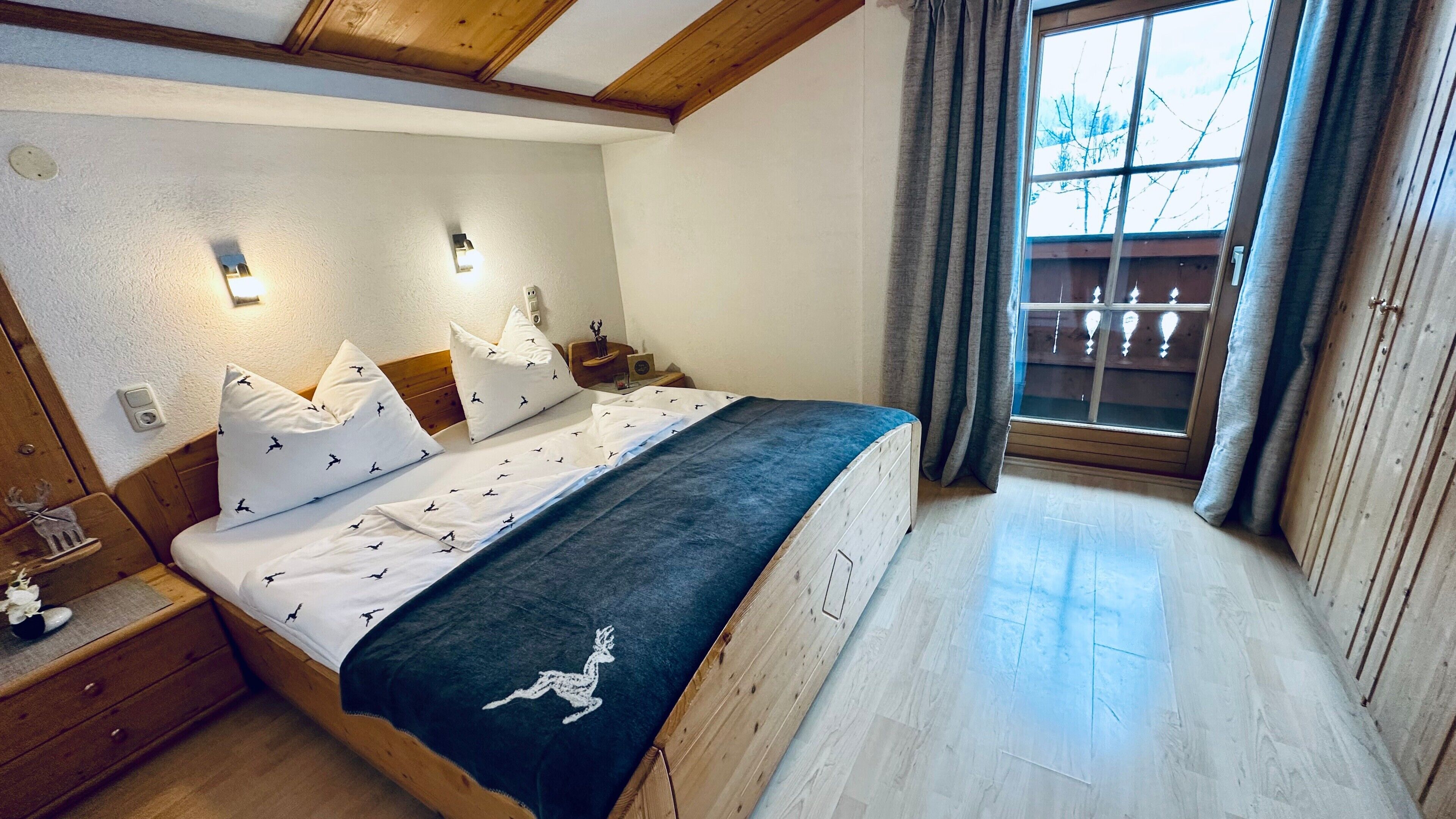 Ein Schlafzimmer mit einem hölzernen Bett, weißer Bettwäsche, einem Nachttisch, Vorhängen an einem Fenster und Blick ins Freie.