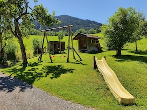 Une aire de jeux avec des balançoires et un toboggan, une maison en bois et un champ d’herbe avec des arbres et des montagnes en arrière-plan.