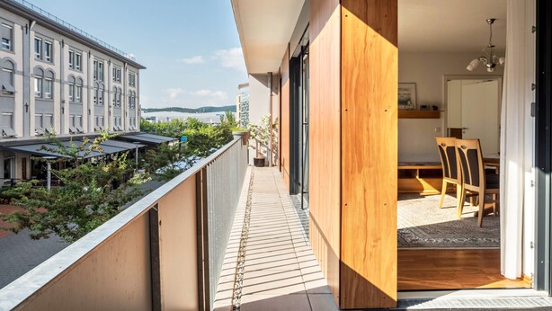 Un balcon donnant sur un immeuble, un coin repas avec des chaises et une table, et une porte en bois menant à l’intérieur.