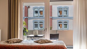 Une chambre à coucher avec un lit, deux oreillers, une fenêtre, une table avec une chaise, et une vue sur un bâtiment à l’extérieur.