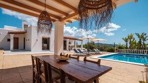 Una mesa de madera con sillas, una lámpara colgante de mimbre, una piscina y un edificio blanco con tejado de baldosas.