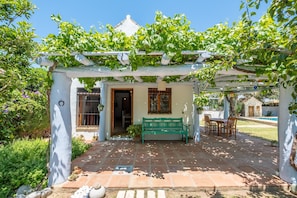 Un patio cubierto con un banco, una mesa y sillas, rodeado de vegetación y una piscina.