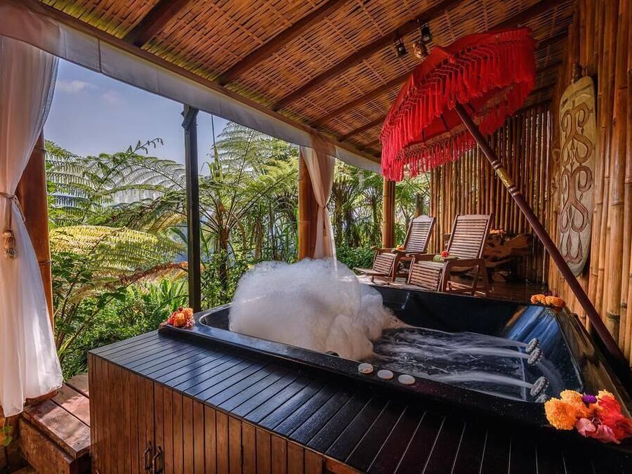 A hot tub with a wooden deck, surrounded by bamboo walls, with chairs and a view of tropical vegetation.