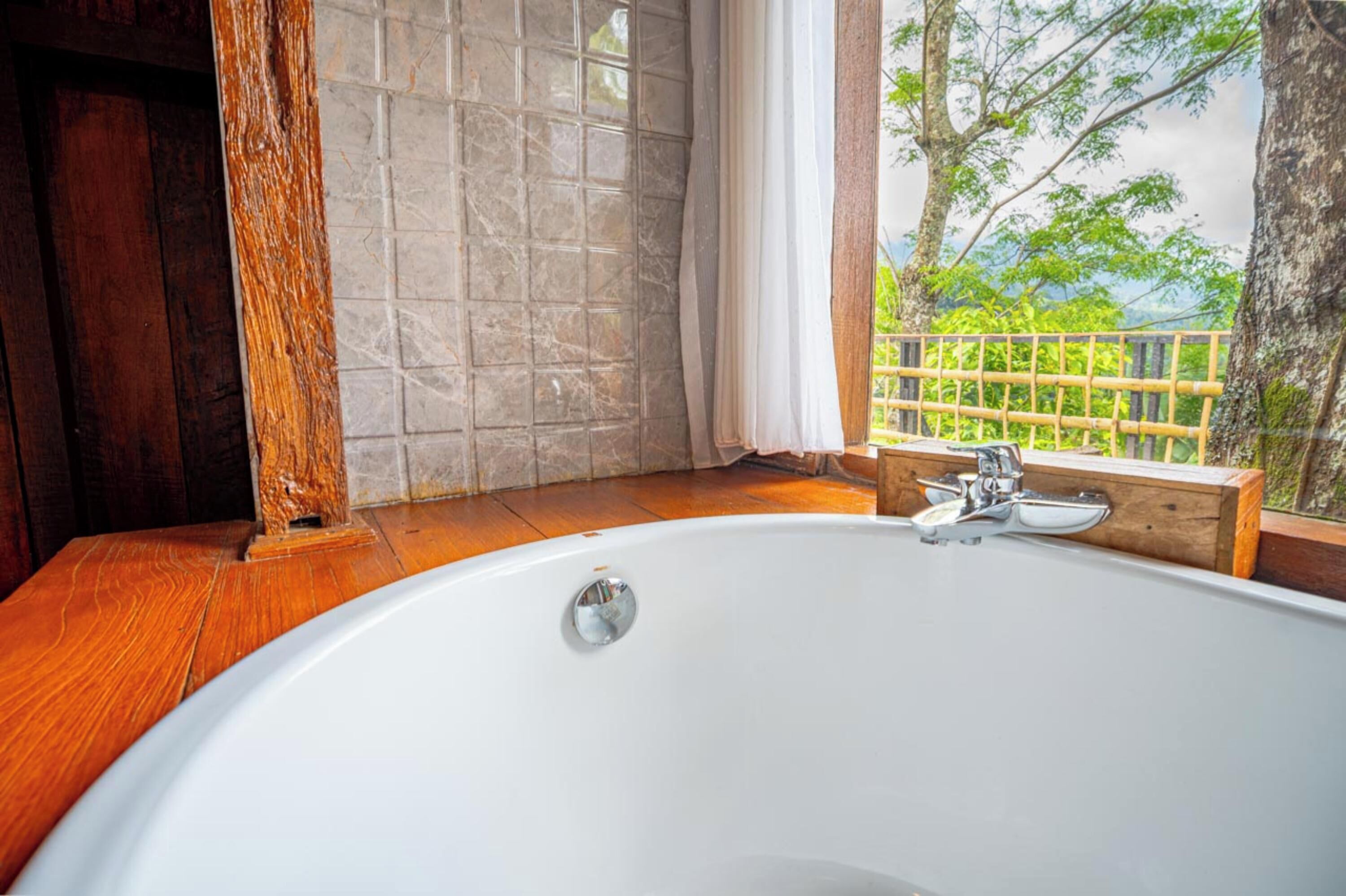 A bathroom with a large bathtub, wooden flooring, a window with a view of greenery, and a wooden bench.